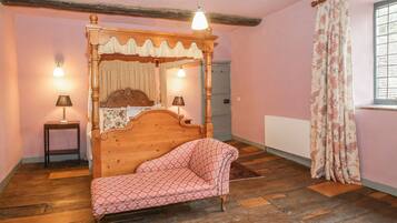 A bedroom with a four-poster bed, two bedside lamps, a wooden floor, a window with floral curtains, and a radiator.