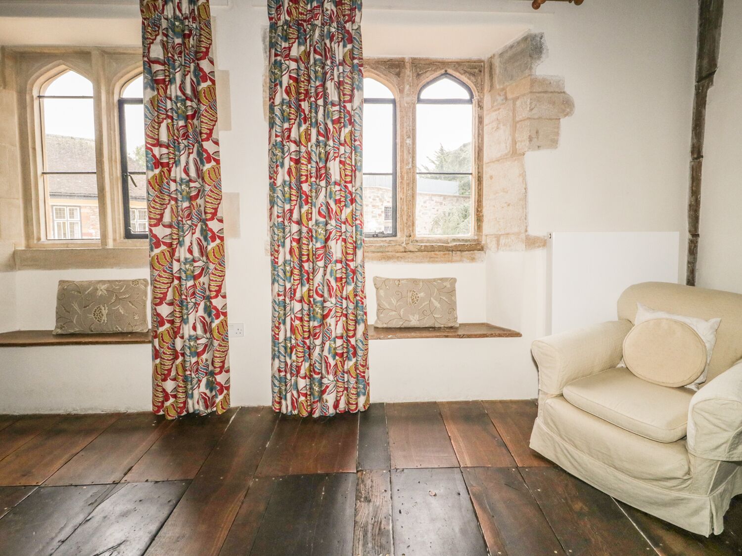 Una habitación con suelo de madera, un sofá color beige y una ventana con vistas a edificios.