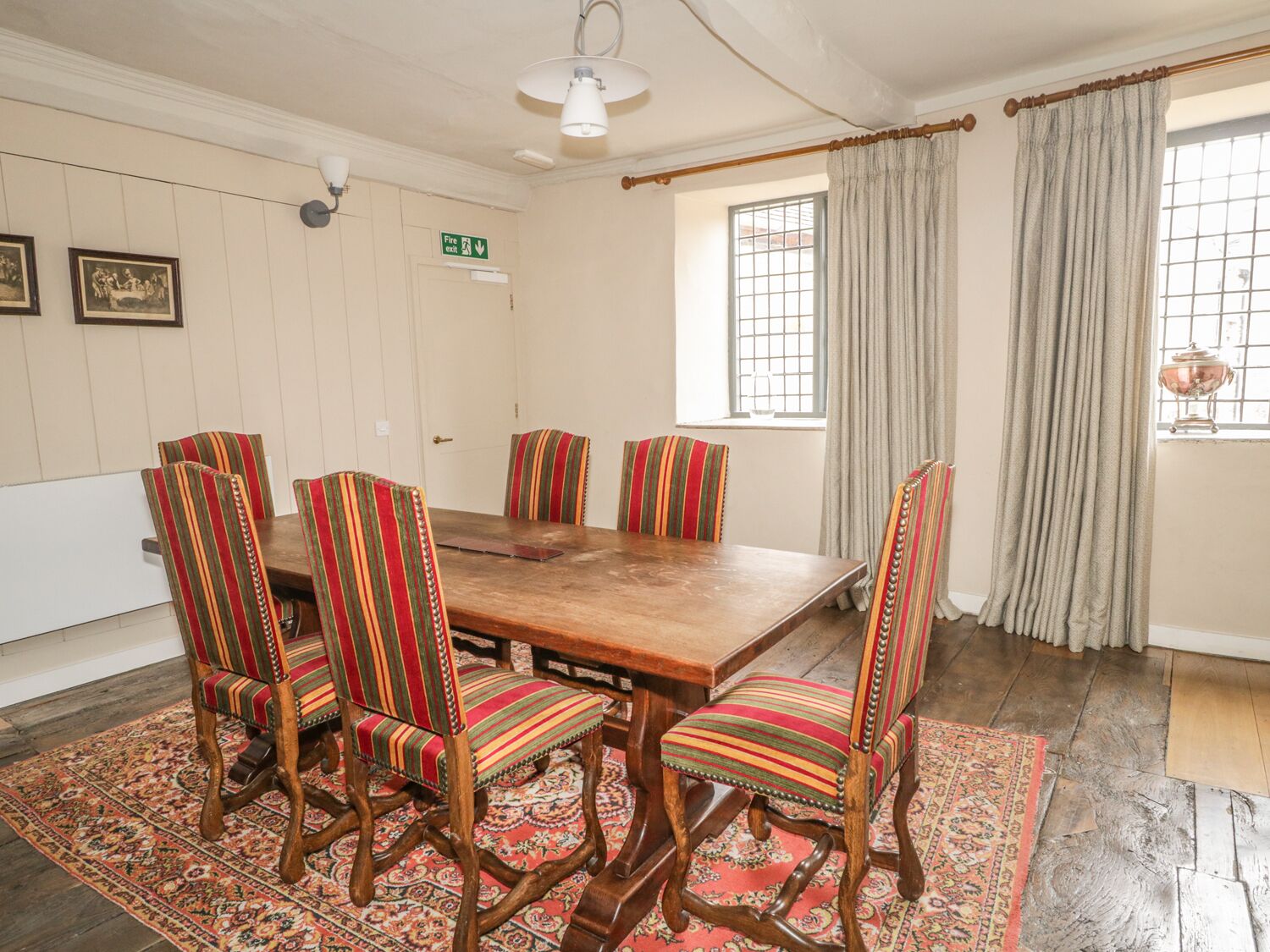 Un comedor con una mesa de madera y seis sillas a rayas, un tapete estampado, un letrero de salida de incendios y una ventana con cortinas.
