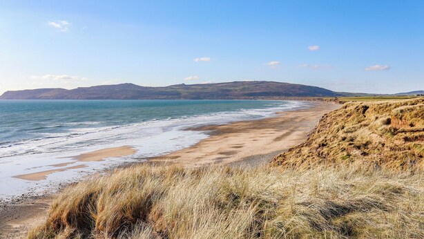 A coastal landscape with a sandy beach, grassy dunes, and distant hills.