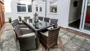 Un patio con una mesa de vidrio y sillas de mimbre, rodeado por un muro blanco con ventanas y una puerta.