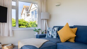 A modern living room with a blue sofa, a yellow pillow, a lamp, and a view of a house through a window. - Grand View (Kingsbridge)