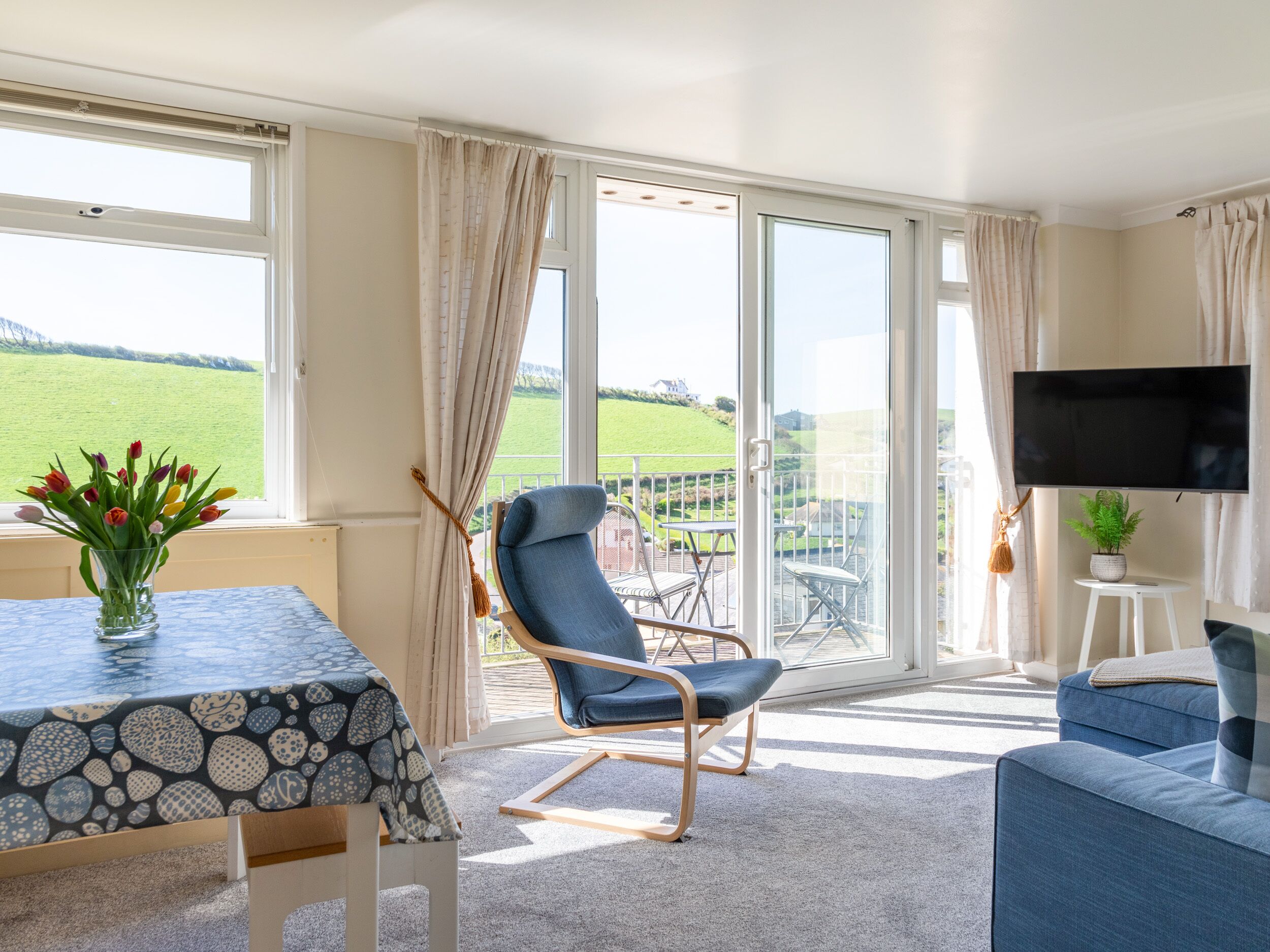 Un salon moderne avec une grande fenêtre, un fauteuil bleu, un canapé, un téléviseur et une table sur laquelle est posé un vase de fleurs.