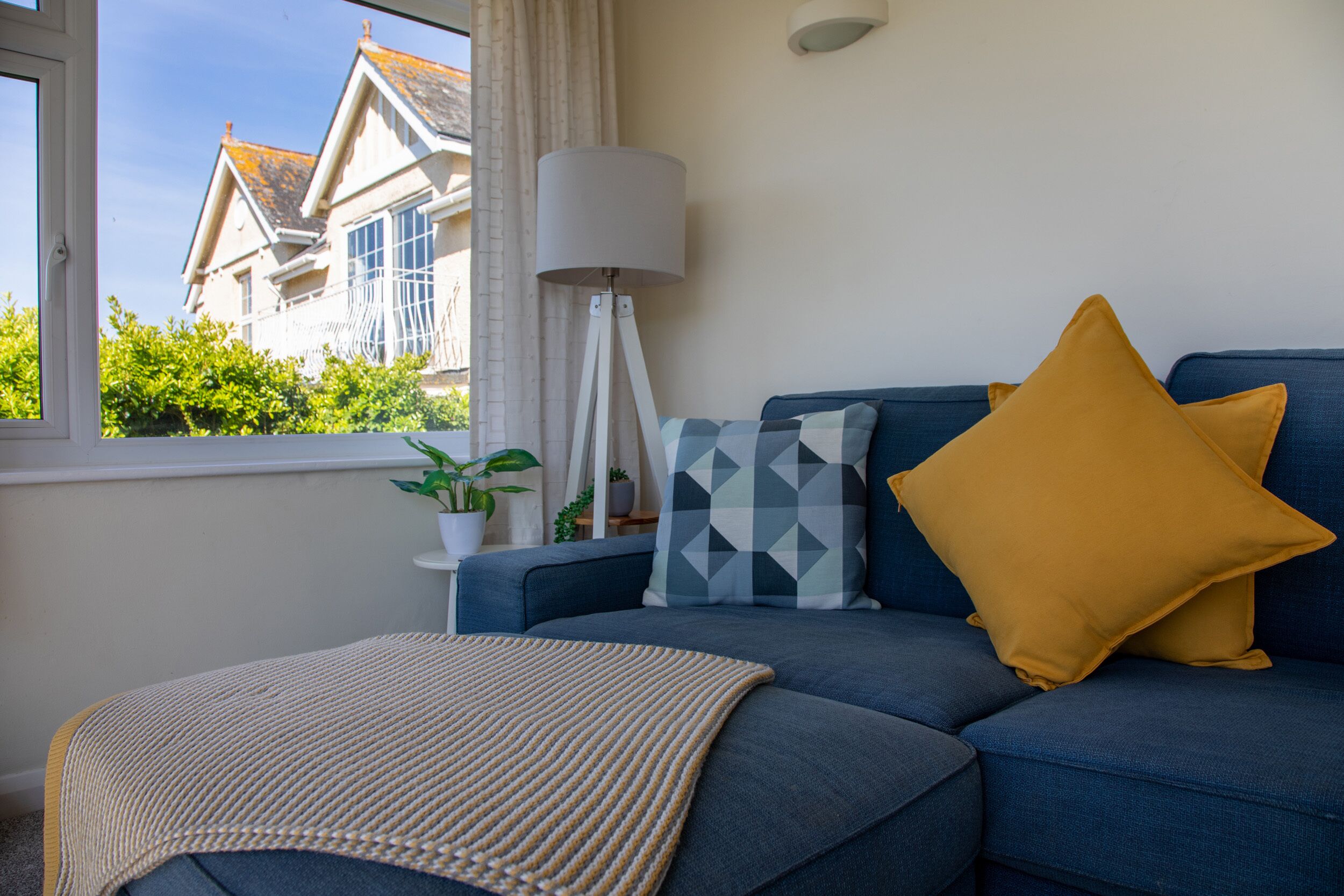 Un salon avec un canapé bleu, un coussin jaune, un coussin à motifs, une lampe sur pied et une vue sur une maison par la fenêtre.