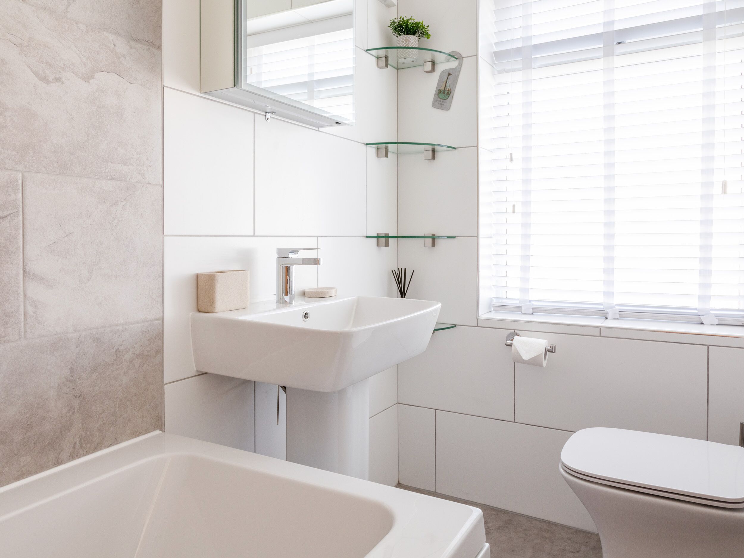 Un baño moderno con bañera, lavabo e inodoro. Hay repisas de vidrio sobre el lavabo y una ventana con persianas.