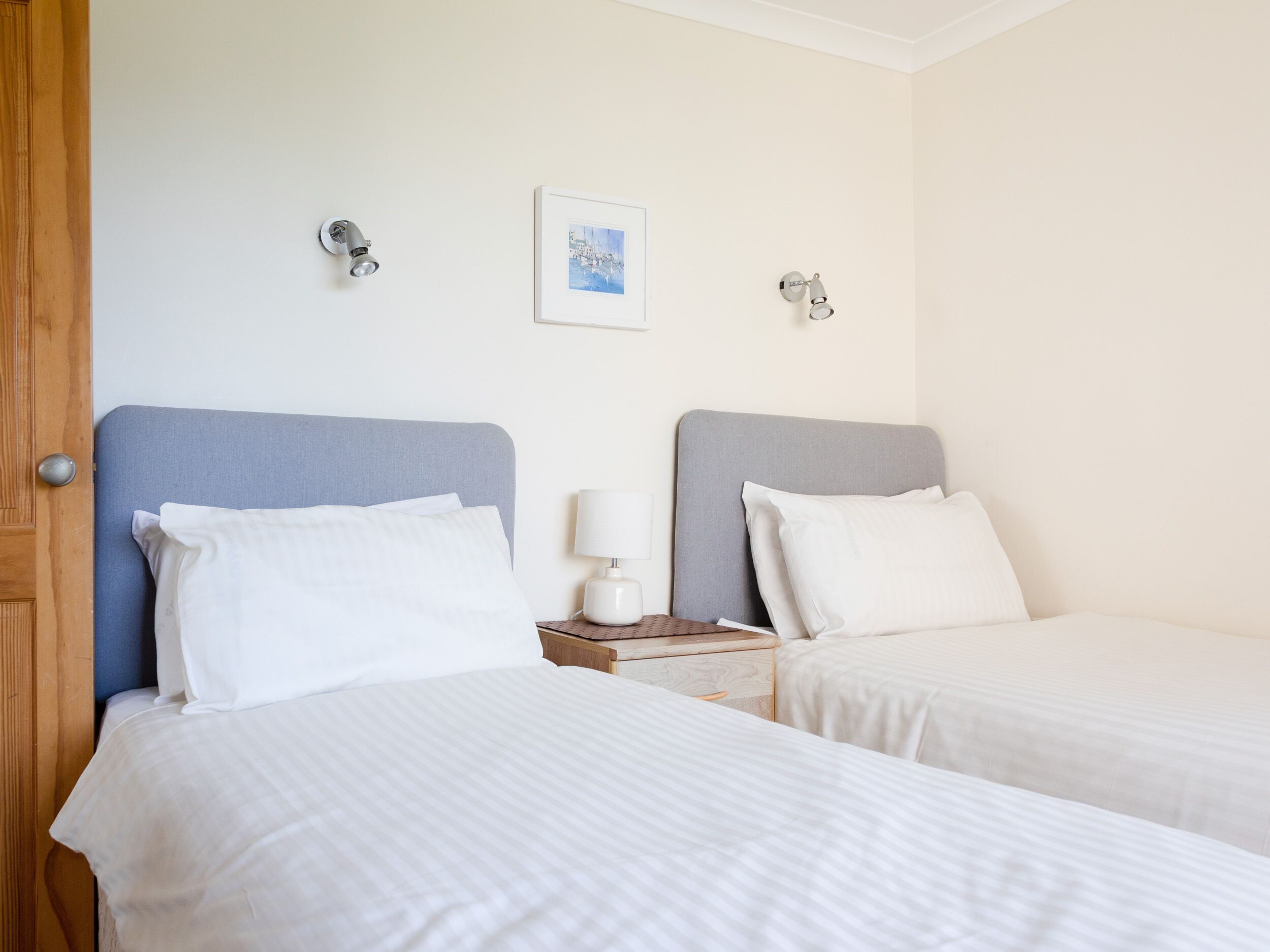 A hotel room with two single beds, a wooden door, a small bedside table with a lamp, and a framed picture on the wall.