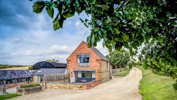 A rural property with a brick house, gravel driveway, and surrounding greenery. - Top Barn (Shipston on Stour)