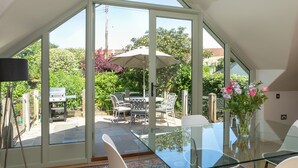 Une pièce spacieuse dotée d’une grande porte-fenêtre coulissante donnant sur un patio extérieur. À l’intérieur, on trouve une table en verre avec des chaises, un vase rempli de fleurs et un ventilateur de plafond.