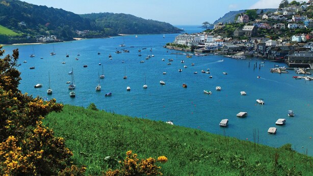 Une ville côtière où des bateaux sont amarrés le long du port, entourée d’une végétation luxuriante et de collines.