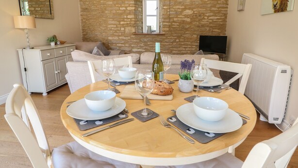 A dining area with a stone wall, a sofa, a table set for four, and a radiator. - Ewecub Barn (Witney)
