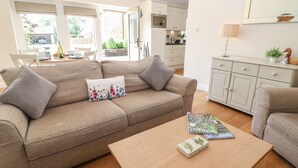 A living room with a beige sofa, a wooden coffee table, and a dining area with a white table and chairs. - Ewecub Barn (Witney)