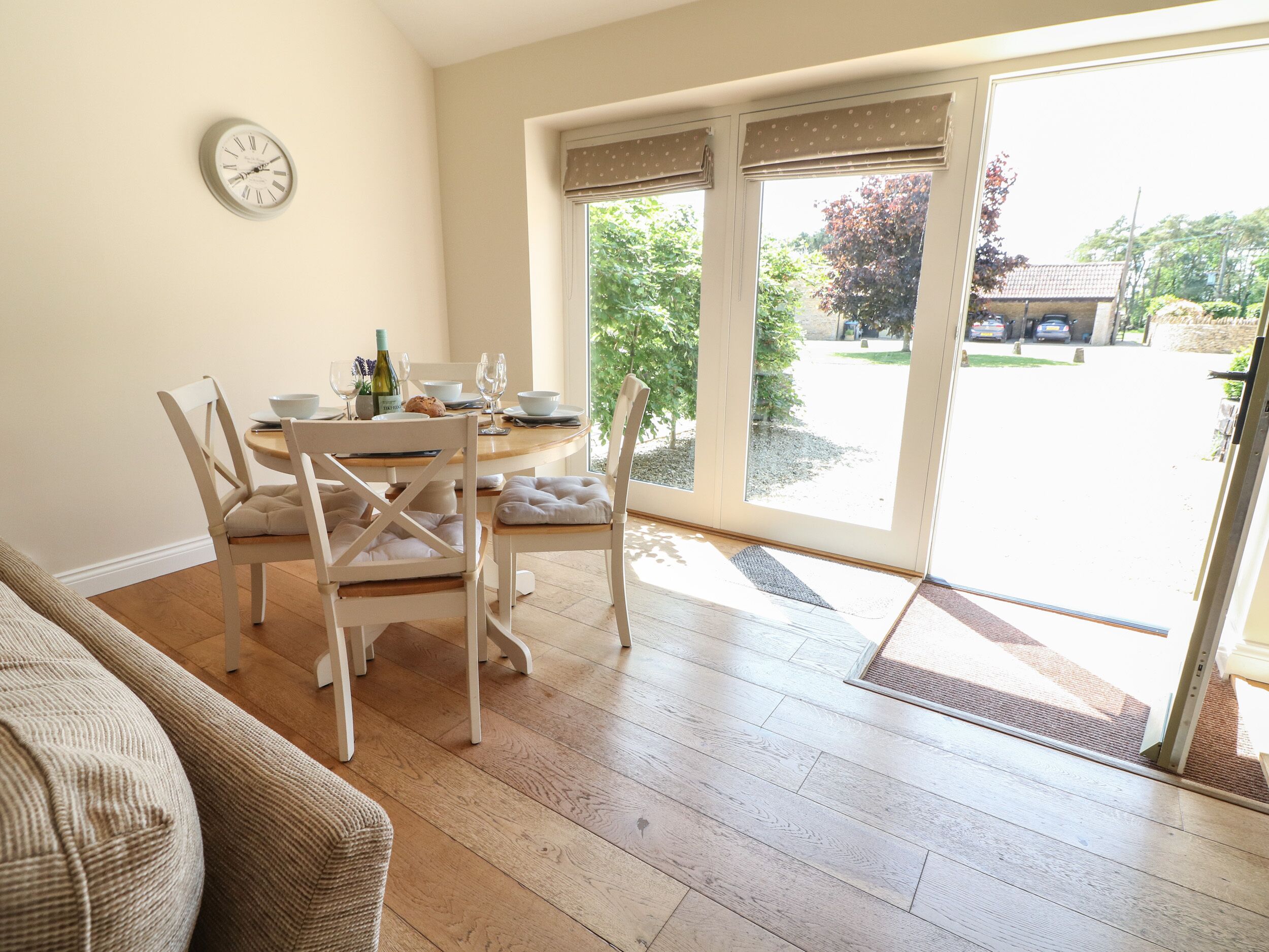 Un coin repas avec une table dressée pour deux personnes, une horloge au mur et une vue sur l’extérieur grâce à de grandes fenêtres.