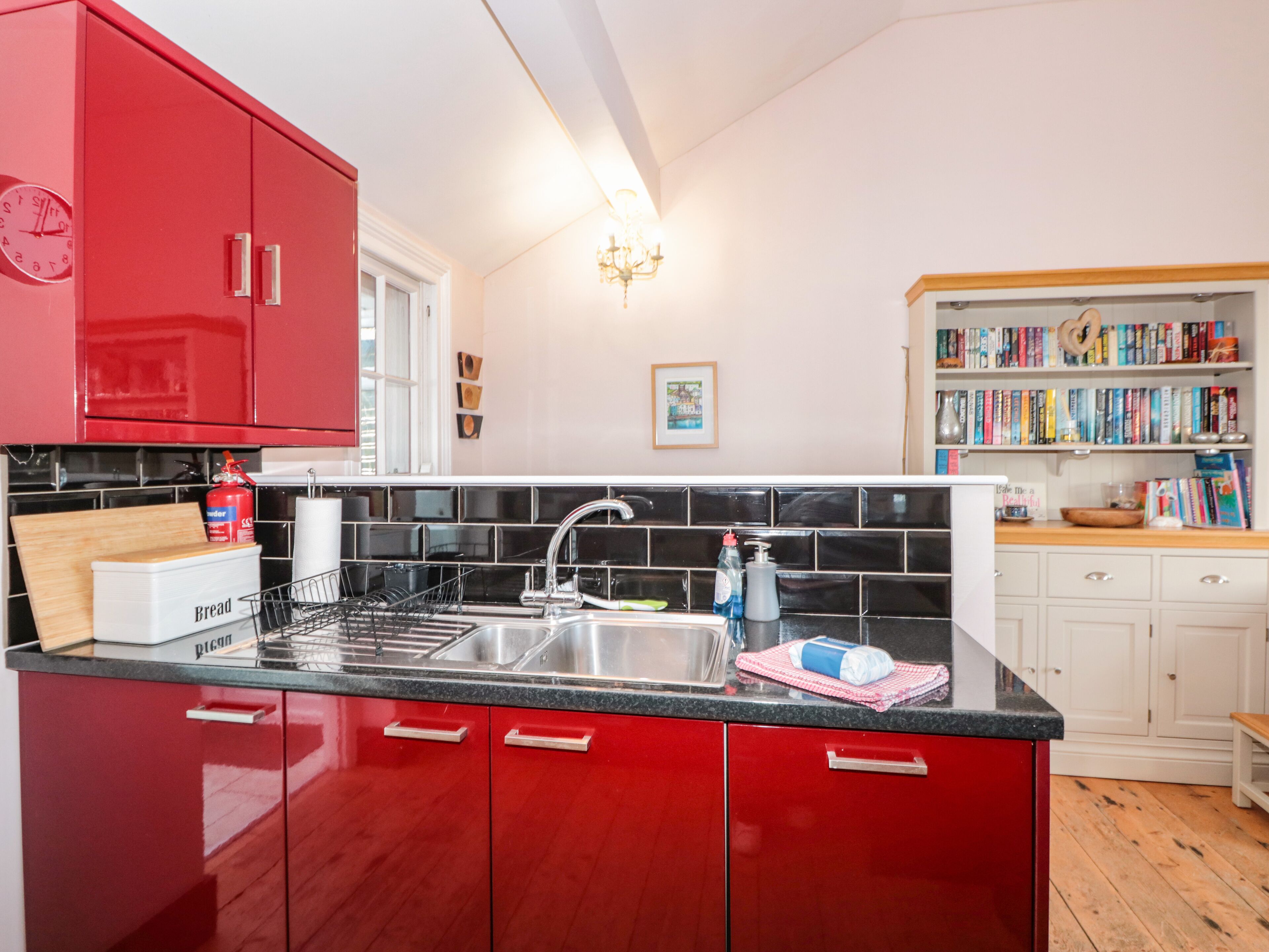 A kitchen with red cabinets, a black countertop, and a sink. There is a bookshelf with books and a framed picture on the wall.