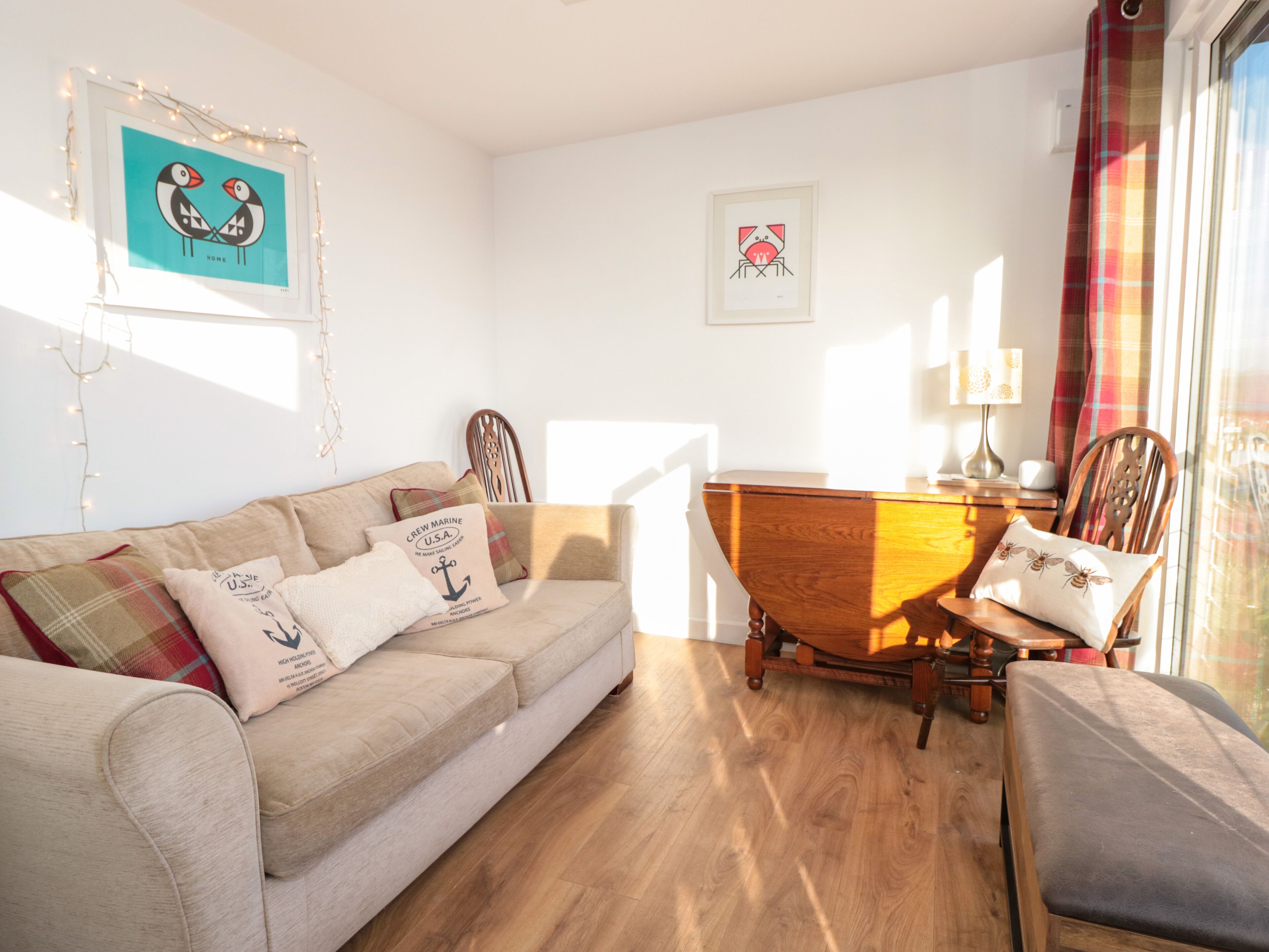 A cozy living room with a wooden floor, a beige sofa, a wooden coffee table, and a chair. There are two framed artworks on the wall and a window with curtains.