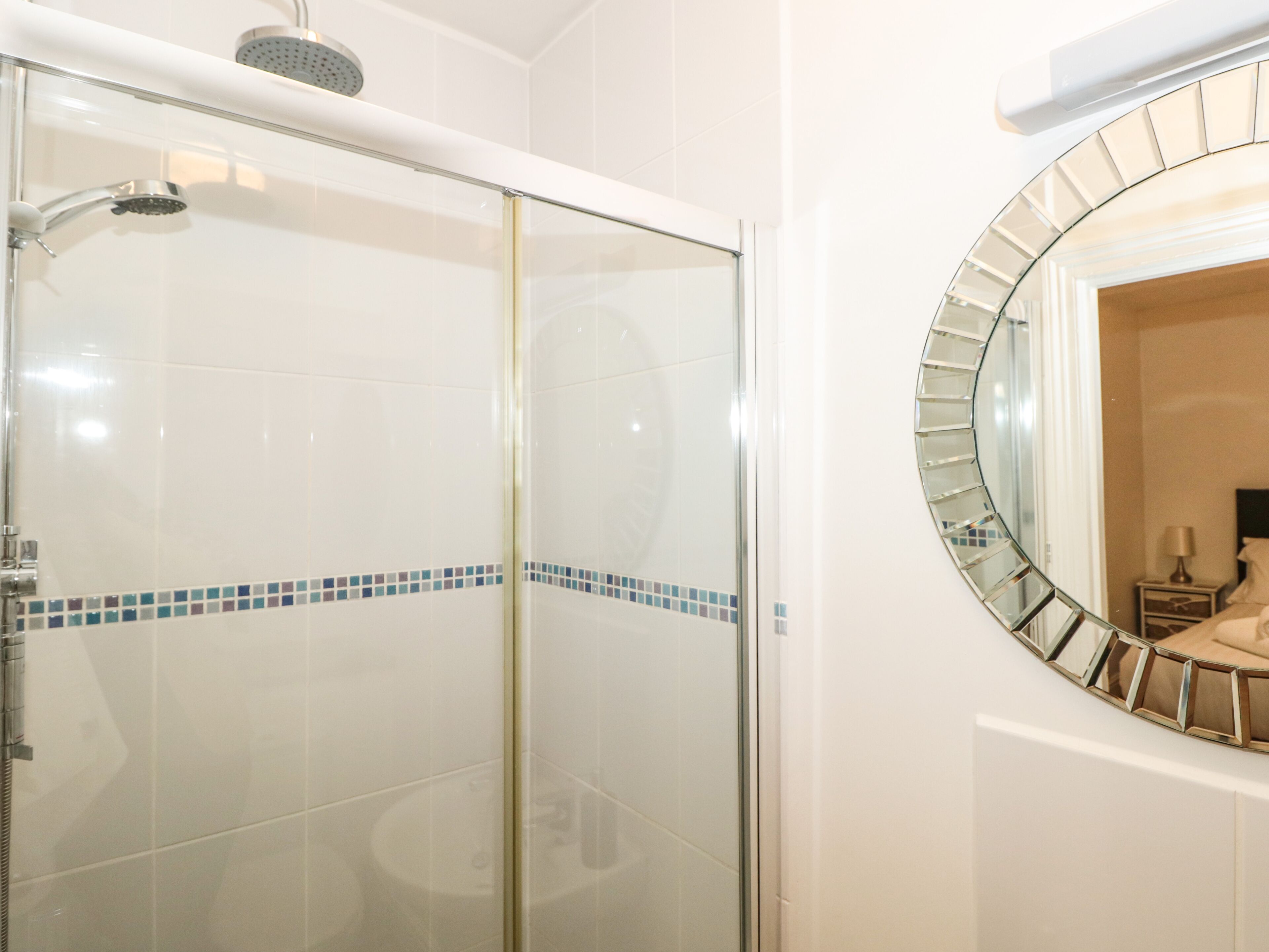 A bathroom with a glass-enclosed shower, a tiled floor, and a round mirror with a metallic frame.