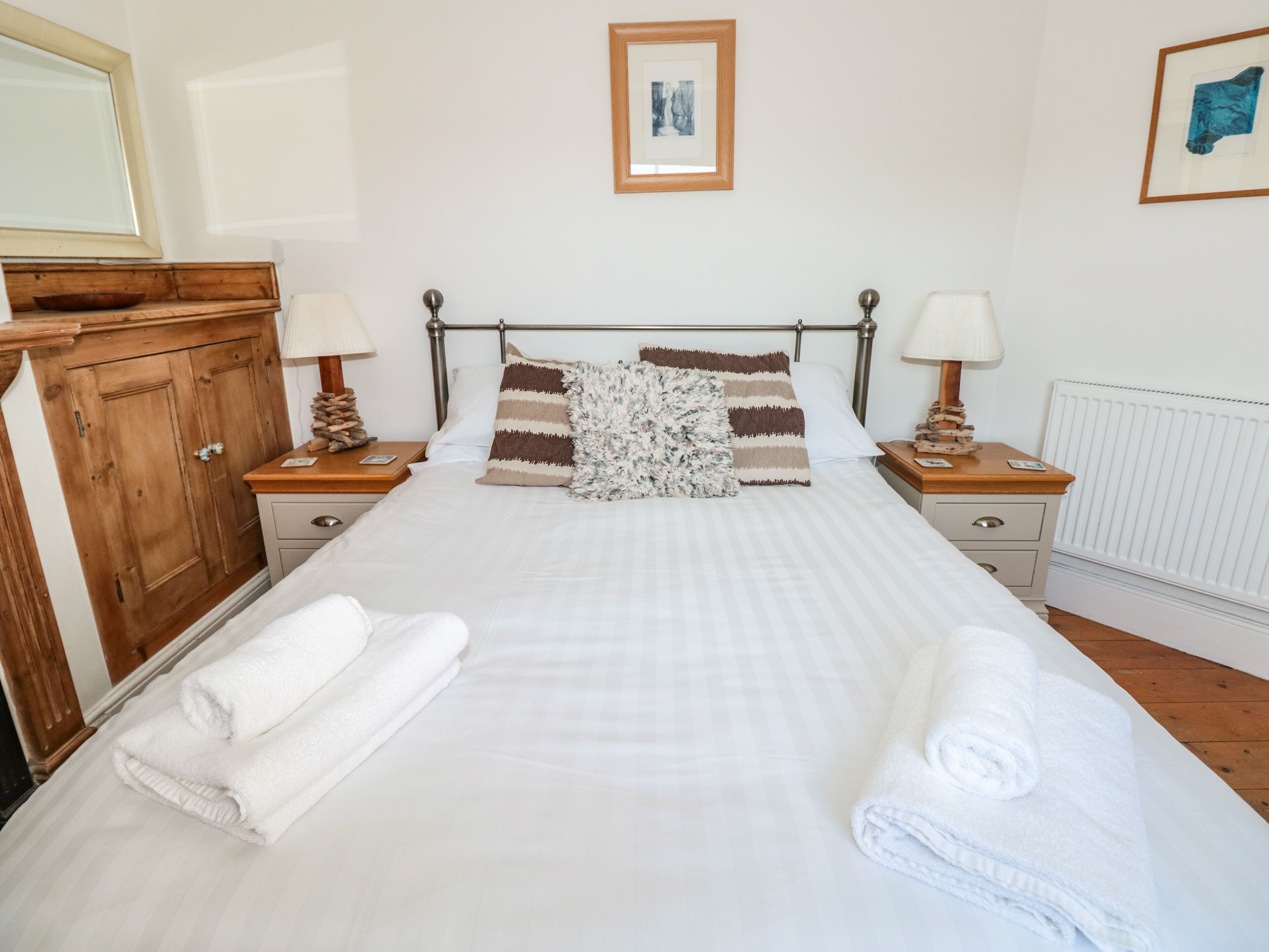 A neatly made bed with white linens and two rolled towels, a wooden nightstand with a lamp, a framed picture on the wall, and a wooden wardrobe.