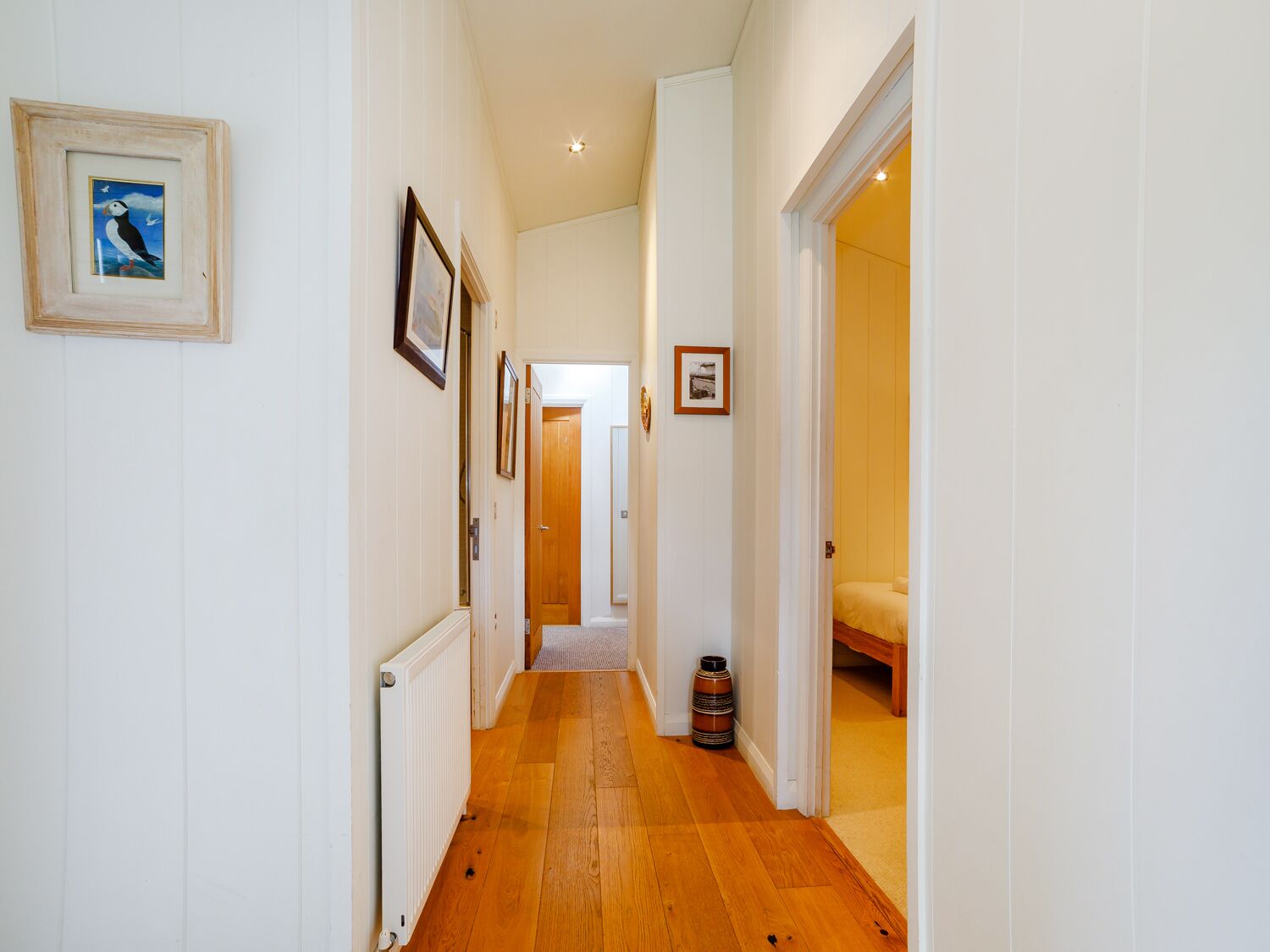 A narrow hallway with wooden flooring, white walls, and framed pictures.