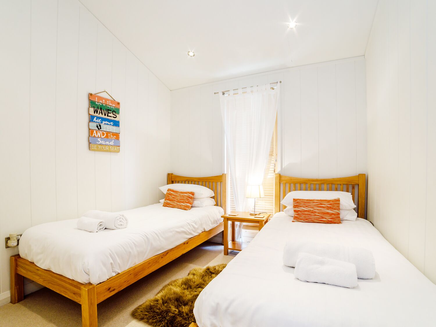 A hotel room with two wooden beds, white bedding, a small wooden nightstand, a fluffy rug, and a sign on the wall.