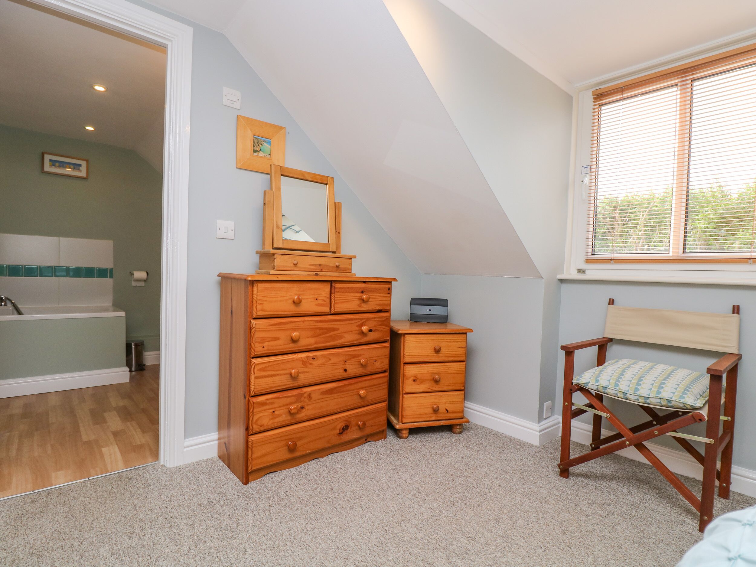 A bedroom with a wooden dresser, a small desk, a chair, and a window with blinds.
