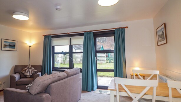A modern living room with a sofa, a dining table, and a chair. There are curtains on the windows, and a lamp is on the floor. - Church House (Achnasheen)