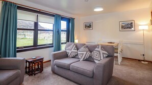 A living room with a grey sofa, a wooden side table, and a window with curtains. There are two wall-mounted lamps and framed pictures on the wall. - Church House (Achnasheen)