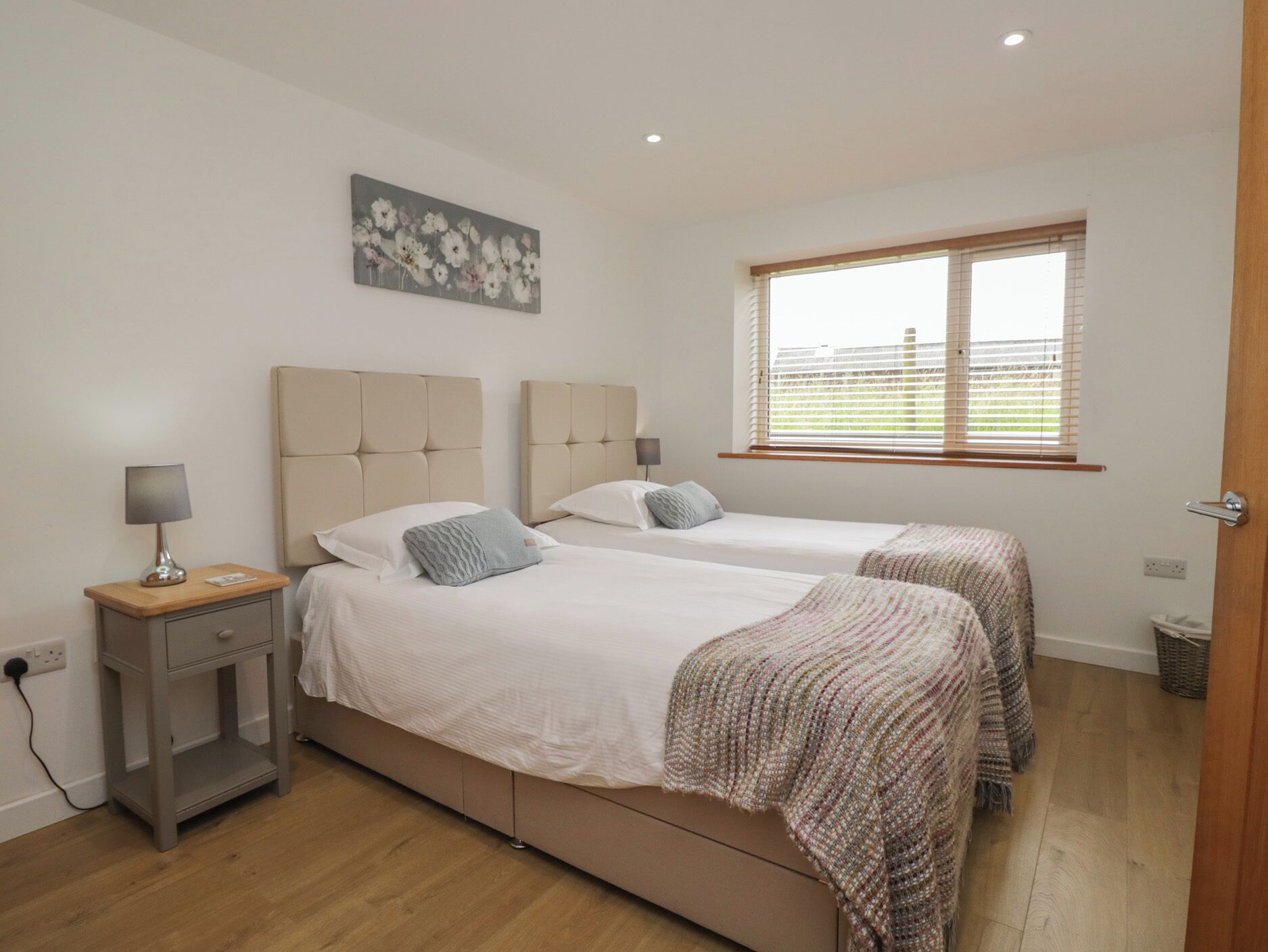 A bedroom with two beds, a bedside table with a lamp, a painting on the wall, and a window with blinds.