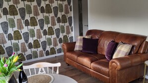 A brown leather sofa with multiple cushions, a round white table with woven placemats, a white chair, and a wall with a tree pattern. - Yew Fold (Kirkby-in-Furness)