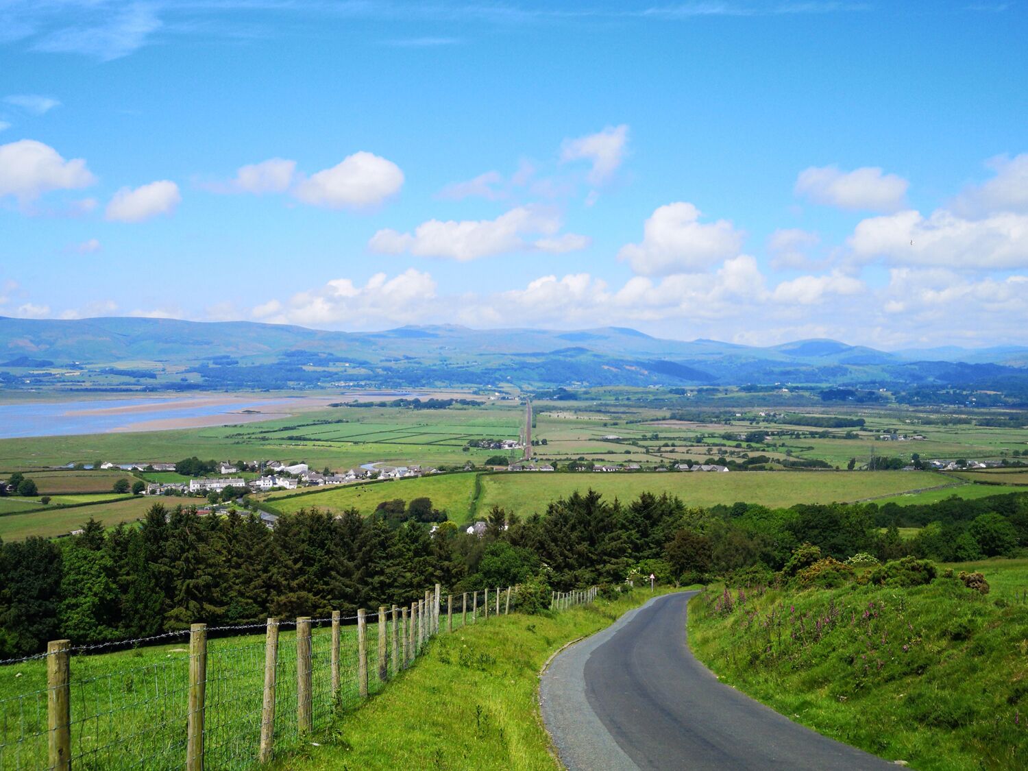 A winding road with a fence on one side and a view of a rural landscape with fields, houses, and mountains in the distance.
