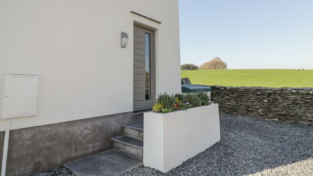 A modern building with a white exterior, a grey door, and a satellite dish. There is a small garden with plants and a stone wall in the foreground. - Yew Fold (Kirkby-in-Furness)