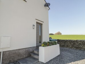 A modern building with a white exterior, a grey door, and a satellite dish. There is a small garden with plants and a stone wall in the foreground. - Yew Fold (Kirkby-in-Furness)
