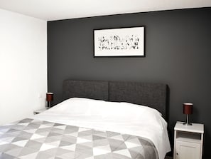 Un dormitorio moderno con cabecera gris, colcha de estampado geométrico, cuadro abstracto enmarcado en la pared y dos lámparas de noche.