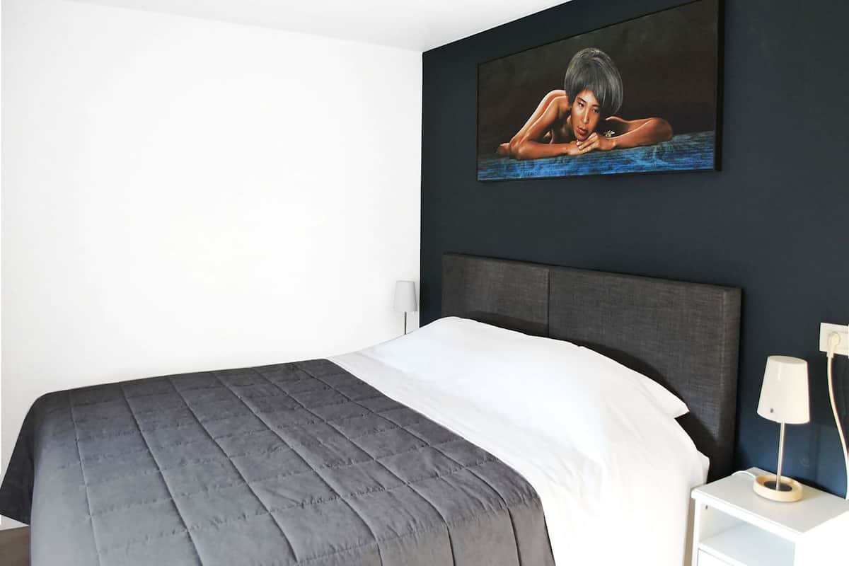 Een moderne slaapkamer met een groot bed, een donker hoofdeinde, een nachtkastje met een lamp en een schilderij aan de muur.
