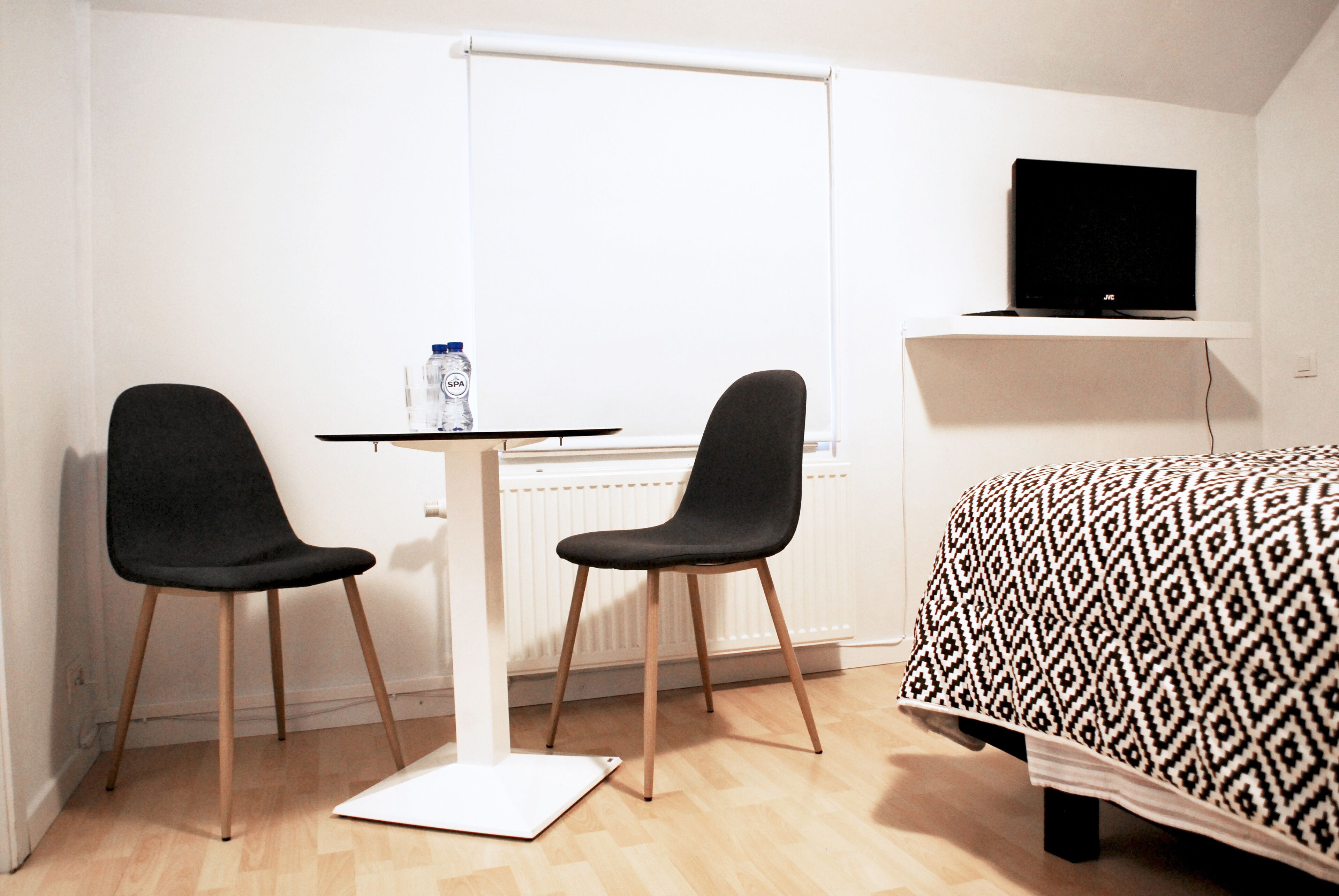 A small room with a bed, a desk with two chairs, a water bottle, and a TV on a shelf.