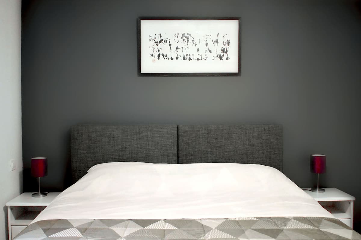 Een moderne slaapkamer met een grijs hoofdeinde, een witte spreidek en een deken met een geometrisch patroon. Er staan twee nachtkastjes met rode lampen.