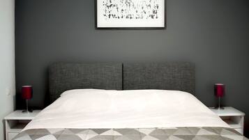 Un dormitorio moderno con cabecera gris, colcha blanca y manta de estampado geométrico. Hay dos mesitas de noche con lámparas rojas.