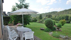Een houten terras met een tafel, stoelen en een parasol, met uitzicht op een groene tuin met bomen en rotsen.