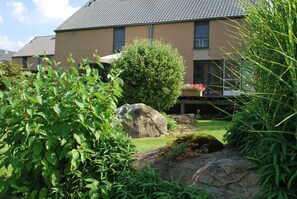 Un immeuble résidentiel à la façade en briques, doté d’un toit en tuiles et d’un jardin bien entretenu, agrémenté d’un grand rocher, de quelques arbustes et d’un petit étang.
