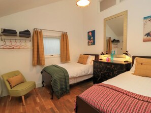 A bedroom with two beds, a chair, a dresser, a window with curtains, a mirror, and a wall-mounted shelf with hangers.