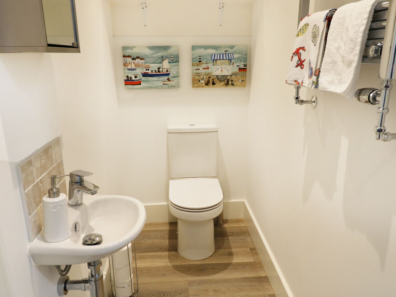Ein kleines Badezimmer mit Toilette, Waschbecken und Handtuchhalter. Zwei an der Wand hängende Bilder zeigen Meeresansichten.