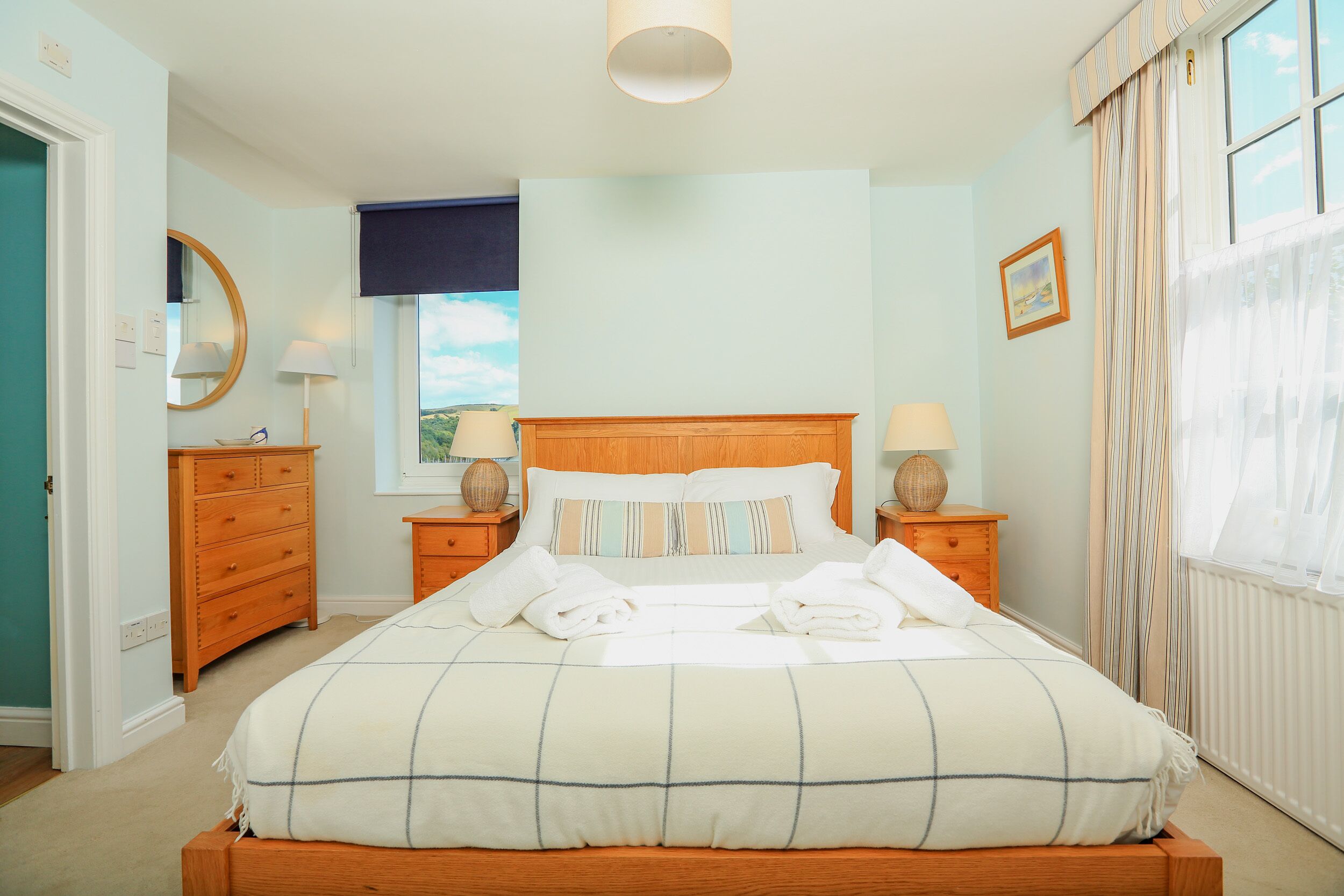 Een net opgemaakt bed met een gestreept dekbed, twee nachtkastjes met lampen, een houten dressoir, een raam met uitzicht en een ingelijste foto aan de muur.