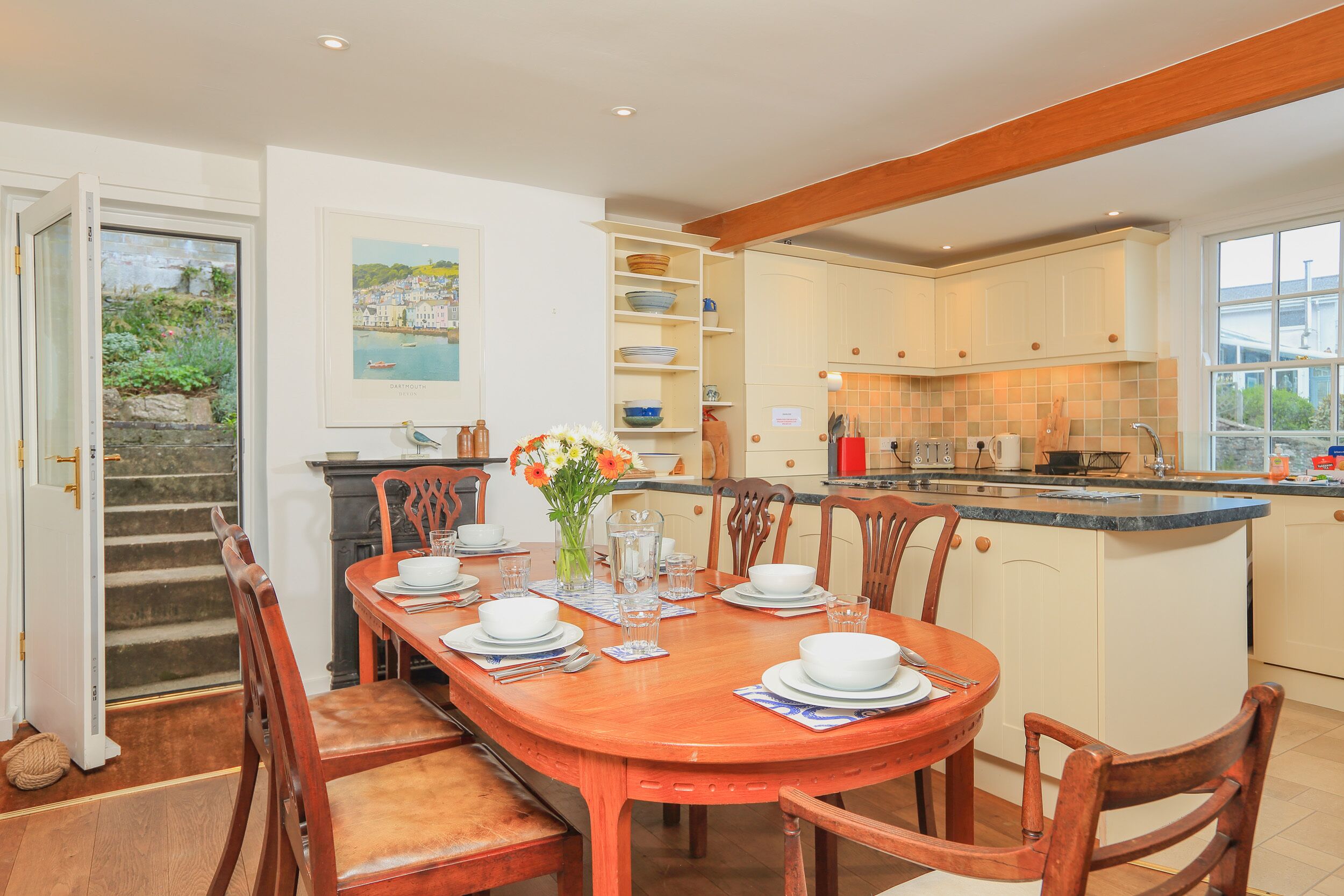 A dining area with a wooden table set for four, surrounded by chairs. A kitchen with white cabinets and a blue countertop is visible in the background.