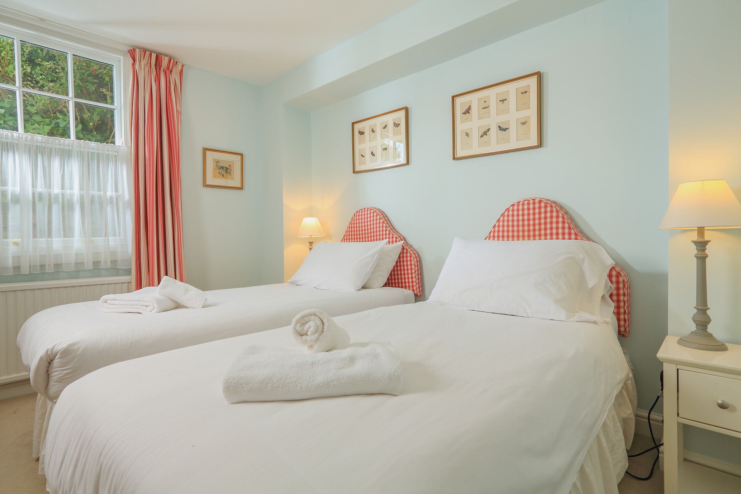 Une chambre d’hôtel avec deux lits, chacun doté de draps blancs et d’une tête de lit à carreaux rouges. On y trouve des tableaux encadrés au mur, une fenêtre avec des rideaux légers et une lampe de chevet.