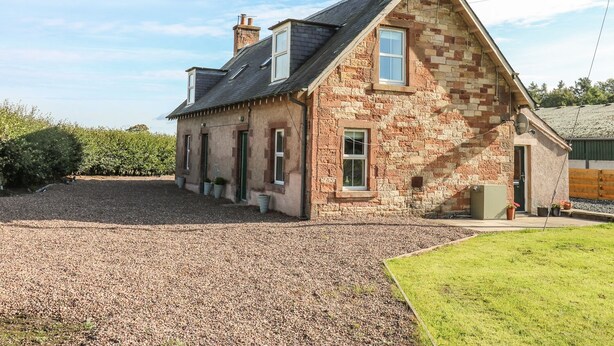 Une maison en pierre avec un toit en pente, une allée en gravier et une petite pelouse.