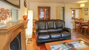 A living room with a black leather sofa, wooden dining table, and chairs. There is a fireplace with a white mantel, a mirror, and a vase with flowers. - Sea Breeze Cottage (Buckie)