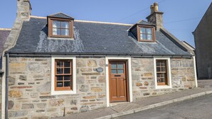 A stone cottage with a brown door, two windows, and a chimney. - Sea Breeze Cottage (Buckie)