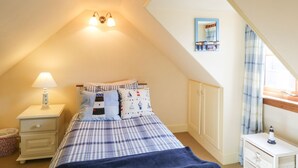 A bedroom with a sloped ceiling, a bed with checkered bedding, a nightstand with a lamp, a wardrobe, and a small table with a lamp. - Sea Breeze Cottage (Buckie)