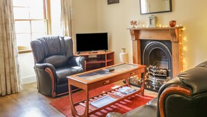 A living room with a leather armchair, a wooden coffee table, a flat-screen TV, a fireplace with lights, and a mirror on the wall. - Sea Breeze Cottage (Buckie)