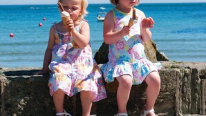 Two children sitting on a stone wall by the sea, wearing floral dresses. - Upper Fernlea (Salcombe)