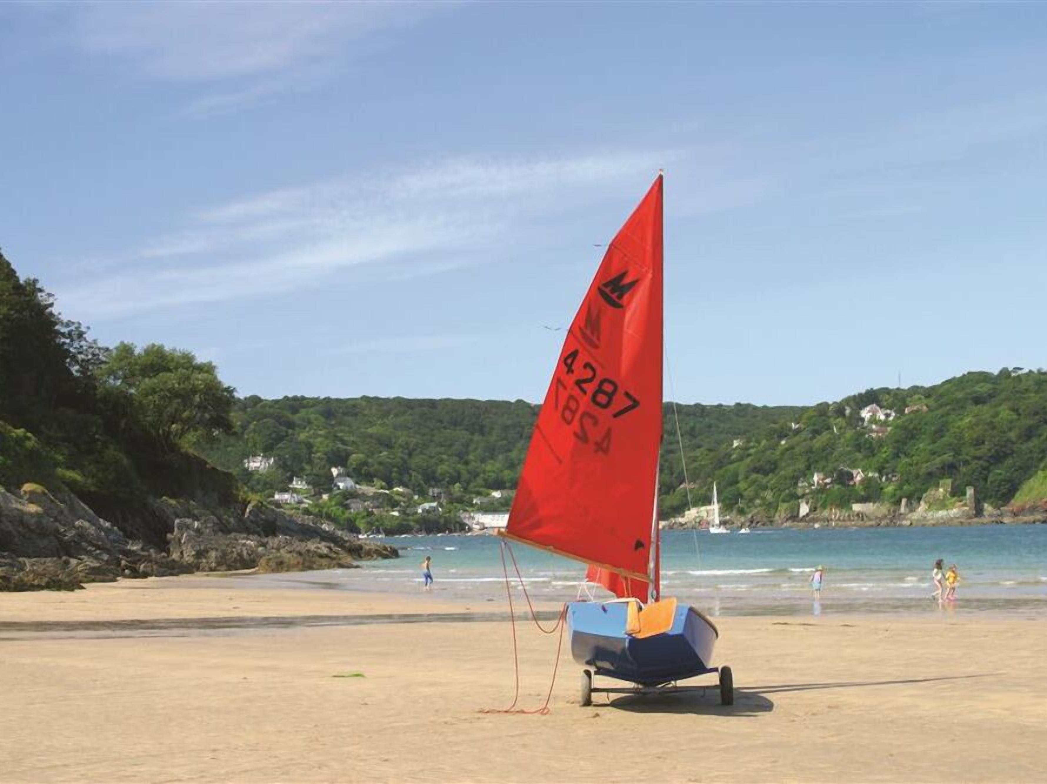 Un velero con vela roja en una playa arenosa, con colinas verdes y cielo despejado al fondo.