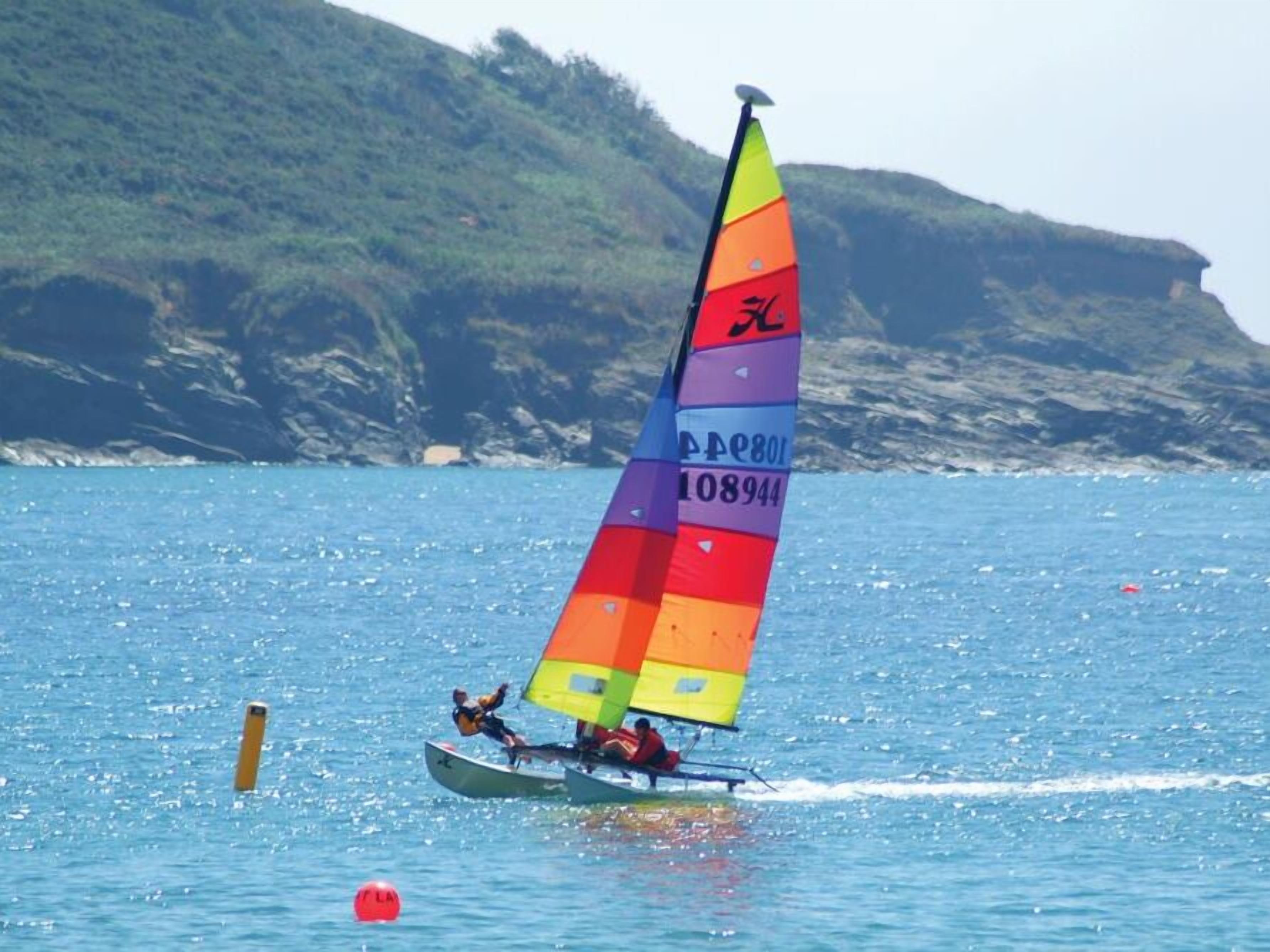 A sailboat with a colorful sail on a body of water with buoys and a rocky coastline in the background.